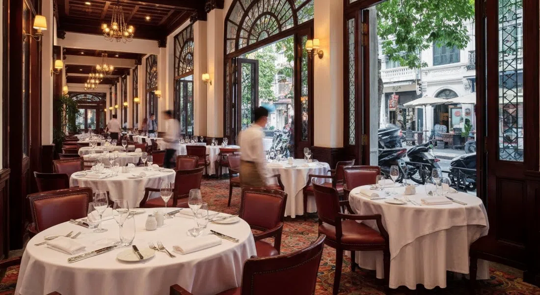 Elegante comedor de Le Beaulieu dentro del Metropole, manteles blancos, cristalería pulida y cálida iluminación colonial que evoca la alta cocina del viejo Hanói